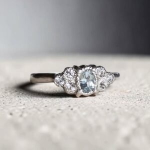 anillo de plata con una piedra aguamarina y 6 zafiros blancos , 3 a cada lado. sobre una superficie blanca de concreto