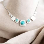 Collar de plata 950 con placas geométricas y turquesas naturales sobre fondo textil neutro.