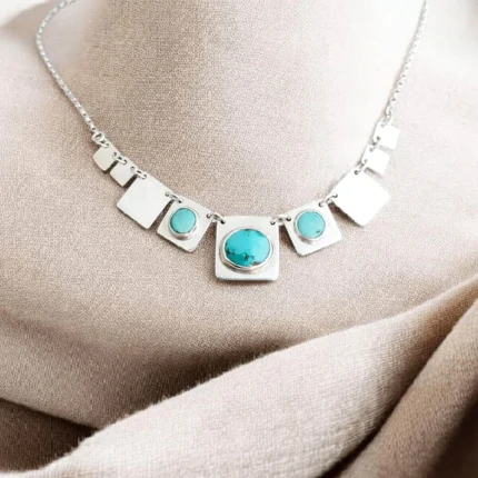 Collar de plata 950 con placas geométricas y turquesas naturales sobre fondo textil neutro.